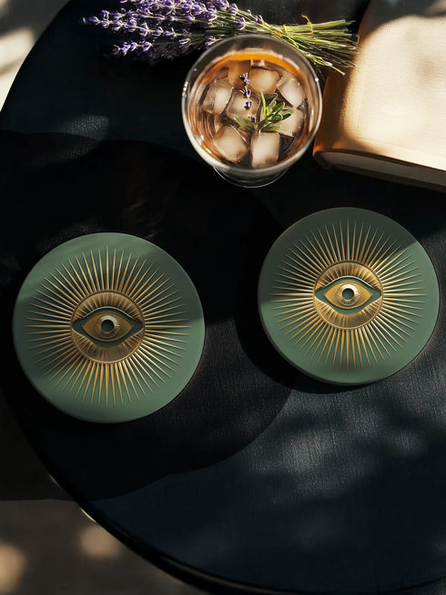 Round ceramic coaster with sage green background featuring a gold Evil Eye symbol in the center with radiating gold sunburst lines extending outward from the eye design.