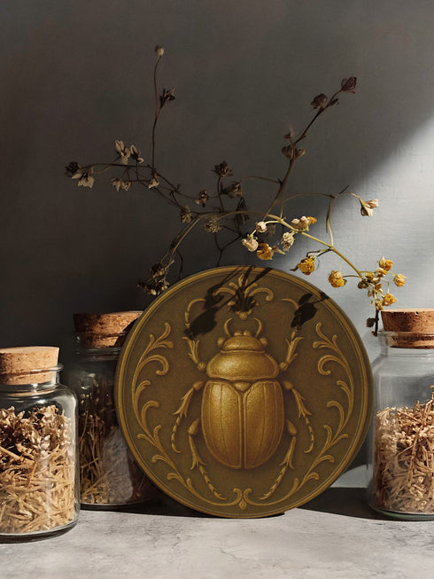 Round ceramic coaster featuring a raised bronze-tone scarab beetle and ornate filigree motif on a matte dark brown glazed surface; 4-inch diameter; backed with cork for non-slip protection; heat-resistant and easy-wipe; perfect for hot or cold beverages in mystical, dark-academic, or antique-inspired interiors.