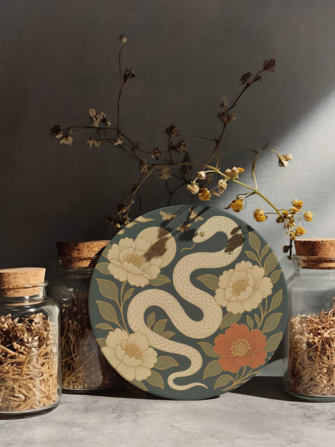 Round ceramic coaster with a chinoiserie-inspired design featuring a white snake among golden peonies, butterflies, and a glowing moon on a dark green background. Surrounded by floral and botanical accents with a soft vintage aesthetic. The coaster has a cork back and measures about 4 inches in diameter. Ideal for mugs, teacups, or wine glasses, this decorative piece suits witchy, victorian, and chinoiserie-inspired decor.