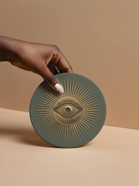 Round ceramic coaster with sage green background featuring a gold Evil Eye symbol in the center with radiating gold sunburst lines extending outward from the eye design.