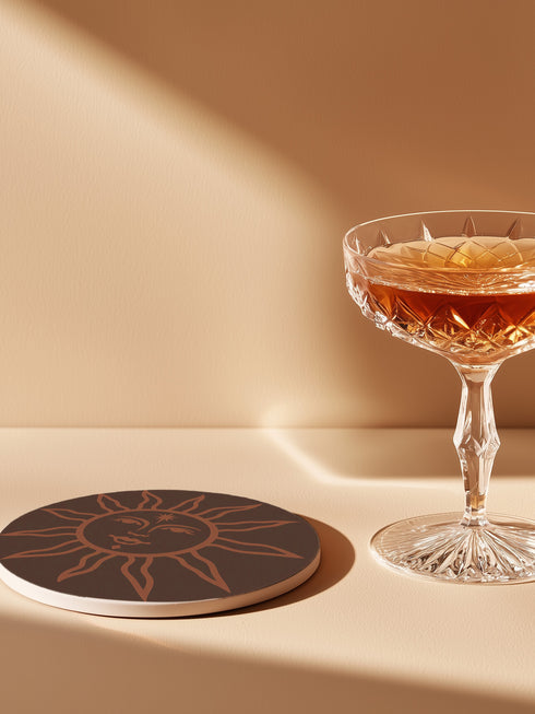 Round ceramic coaster in deep black featuring an intricate burnt-orange sun illustration with a serene, symmetrical face, radiating curved rays, and a delicate star detail on the forehead. The smooth, matte surface enhances the contrast between the dark background and the warm-toned design, creating a celestial, vintage-inspired aesthetic.