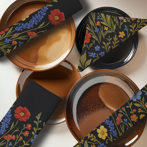 Four-piece cloth napkin set featuring a richly embroidered-style wildflower motif on a deep black background. Includes yellow, red, blue, and orange florals with intricate green stems and leaves. Each napkin is printed on one side and measures 18 x 18 inches, made from smooth polyester fabric with white backing.