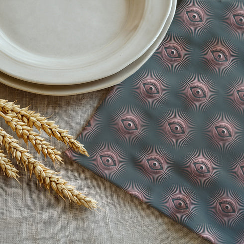 Set of four teal-colored dining napkins featuring a captivating pattern of protective evil eye symbols in dusty rose pink with radiating white lines. Each napkin displays numerous evil eyes arranged in a repeating pattern across the entire surface. The eyes have a vintage, almost Art Deco quality with their symmetrical rays emanating outward like sunbursts. The teal background creates a rich, moody contrast against the rose-pink eyes, giving these napkins a sophisticated, mystical appearance.