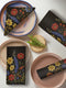 Four-piece cloth napkin set featuring a richly embroidered-style wildflower motif on a deep black background. Includes yellow, red, blue, and orange florals with intricate green stems and leaves. Each napkin is printed on one side and measures 18 x 18 inches, made from smooth polyester fabric with white backing.