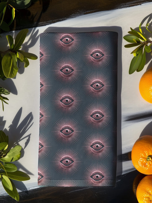 Set of four teal-colored dining napkins featuring a captivating pattern of protective evil eye symbols in dusty rose pink with radiating white lines. Each napkin displays numerous evil eyes arranged in a repeating pattern across the entire surface. The eyes have a vintage, almost Art Deco quality with their symmetrical rays emanating outward like sunbursts. The teal background creates a rich, moody contrast against the rose-pink eyes, giving these napkins a sophisticated, mystical appearance.