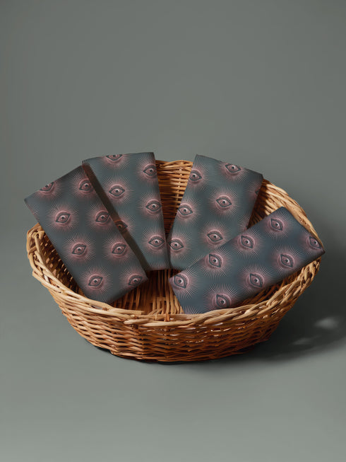 Set of four teal-colored dining napkins featuring a captivating pattern of protective evil eye symbols in dusty rose pink with radiating white lines. Each napkin displays numerous evil eyes arranged in a repeating pattern across the entire surface. The eyes have a vintage, almost Art Deco quality with their symmetrical rays emanating outward like sunbursts. The teal background creates a rich, moody contrast against the rose-pink eyes, giving these napkins a sophisticated, mystical appearance.