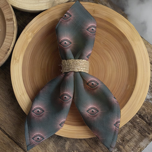 Set of four teal-colored dining napkins featuring a captivating pattern of protective evil eye symbols in dusty rose pink with radiating white lines. Each napkin displays numerous evil eyes arranged in a repeating pattern across the entire surface. The eyes have a vintage, almost Art Deco quality with their symmetrical rays emanating outward like sunbursts. The teal background creates a rich, moody contrast against the rose-pink eyes, giving these napkins a sophisticated, mystical appearance.