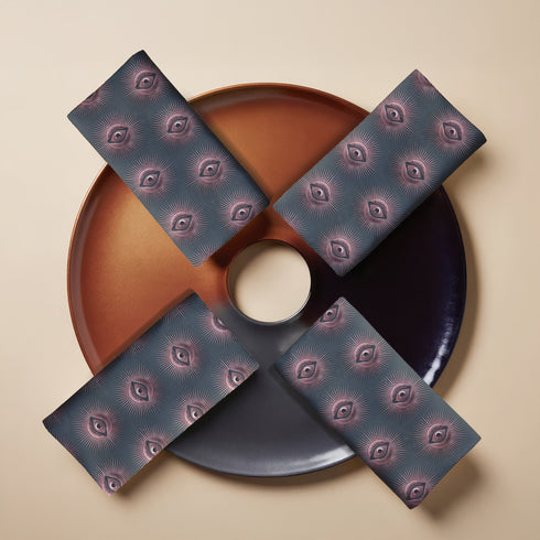 Set of four teal-colored dining napkins featuring a captivating pattern of protective evil eye symbols in dusty rose pink with radiating white lines. Each napkin displays numerous evil eyes arranged in a repeating pattern across the entire surface. The eyes have a vintage, almost Art Deco quality with their symmetrical rays emanating outward like sunbursts. The teal background creates a rich, moody contrast against the rose-pink eyes, giving these napkins a sophisticated, mystical appearance.