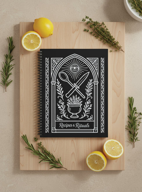 A black spiral-bound hardcover recipe journal lies flat. The cover is matte black with a white line-art design titled "Recipes & Rituals." The art nouveau style illustration features an all-seeing eye under a sunburst, framed by an ornate arch. Below the eye, a crossed spoon and knife are centered above a mortar and pestle filled with herbs. The entire design is bordered by intricate botanical flourishes. The notebook is 8.5 x 11 inches with a wire-o binding, a lay-flat cookbook for kitchen use.