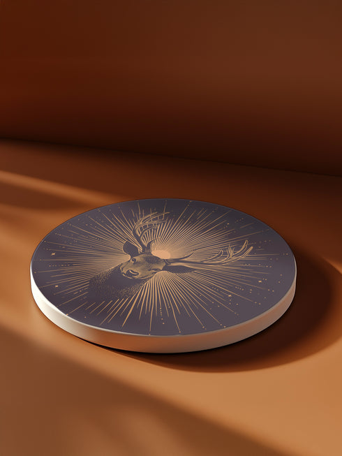 Ceramic coasters in round and square shapes featuring a detailed deer head with branching antlers on a deep navy background. Metallic gold sunburst rays radiate from behind the deer, with scattered stars throughout the design. Each coaster has a protective cork backing.