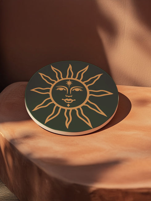 Dark green round ceramic coaster featuring a golden sun illustration with a serene, symmetrical face, delicate curved rays extending outward, and a small star detail on the forehead. The smooth, matte surface enhances the contrast between the deep green background and the warm golden design, giving it a celestial and vintage-inspired aesthetic.