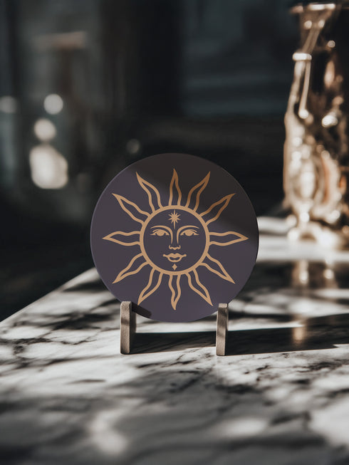 Round ceramic coaster featuring a celestial sun design in warm golden tones against a deep matte black background. The intricate illustration showcases a serene sun face with delicate detailing, surrounded by radiant, curved rays. The smooth, durable surface is complemented by a cork backing for grip and protection, making it both a functional and decorative piece for any space.