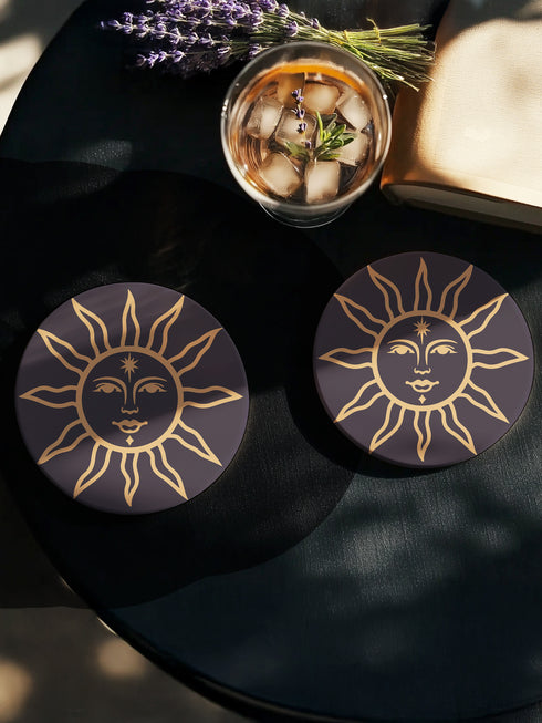 Round ceramic coaster featuring a celestial sun design in warm golden tones against a deep matte black background. The intricate illustration showcases a serene sun face with delicate detailing, surrounded by radiant, curved rays. The smooth, durable surface is complemented by a cork backing for grip and protection, making it both a functional and decorative piece for any space.