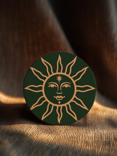 Dark green round ceramic coaster featuring a golden sun illustration with a serene, symmetrical face, delicate curved rays extending outward, and a small star detail on the forehead. The smooth, matte surface enhances the contrast between the deep green background and the warm golden design, giving it a celestial and vintage-inspired aesthetic.