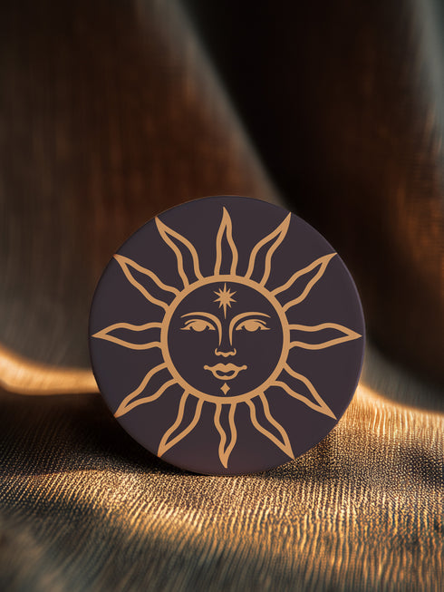 Round ceramic coaster featuring a celestial sun design in warm golden tones against a deep matte black background. The intricate illustration showcases a serene sun face with delicate detailing, surrounded by radiant, curved rays. The smooth, durable surface is complemented by a cork backing for grip and protection, making it both a functional and decorative piece for any space.