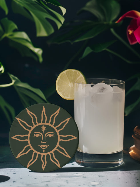 Dark green round ceramic coaster featuring a golden sun illustration with a serene, symmetrical face, delicate curved rays extending outward, and a small star detail on the forehead. The smooth, matte surface enhances the contrast between the deep green background and the warm golden design, giving it a celestial and vintage-inspired aesthetic.
