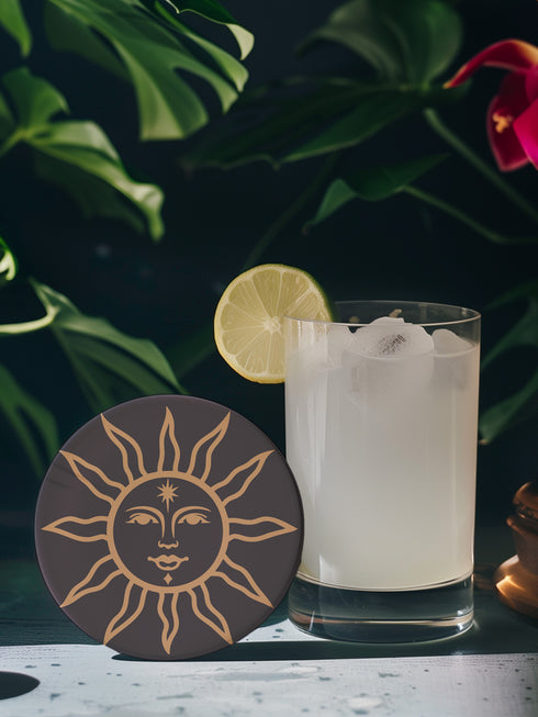 Round ceramic coaster featuring a celestial sun design in warm golden tones against a deep matte black background. The intricate illustration showcases a serene sun face with delicate detailing, surrounded by radiant, curved rays. The smooth, durable surface is complemented by a cork backing for grip and protection, making it both a functional and decorative piece for any space.