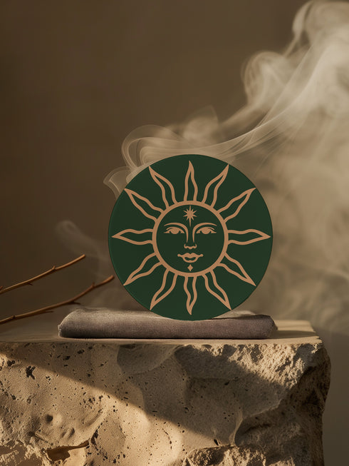 Dark green round ceramic coaster featuring a golden sun illustration with a serene, symmetrical face, delicate curved rays extending outward, and a small star detail on the forehead. The smooth, matte surface enhances the contrast between the deep green background and the warm golden design, giving it a celestial and vintage-inspired aesthetic.