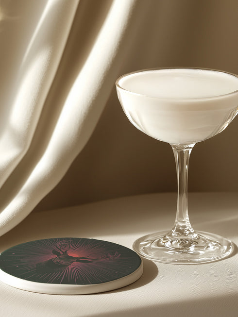 Ceramic coasters in round and square shapes featuring a detailed deer head with majestic antlers against a deep forest green background. A pink-tinged radiant sunburst pattern emanates from behind the deer's head, with delicate stars scattered throughout the design. Cork backing protection.