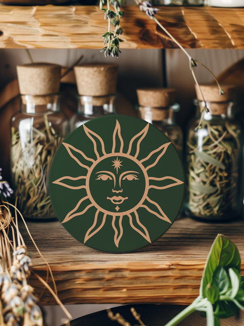 Dark green round ceramic coaster featuring a golden sun illustration with a serene, symmetrical face, delicate curved rays extending outward, and a small star detail on the forehead. The smooth, matte surface enhances the contrast between the deep green background and the warm golden design, giving it a celestial and vintage-inspired aesthetic.