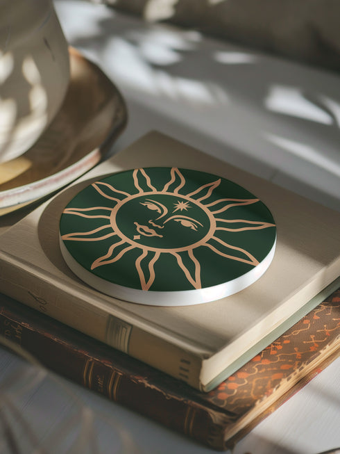 Dark green round ceramic coaster featuring a golden sun illustration with a serene, symmetrical face, delicate curved rays extending outward, and a small star detail on the forehead. The smooth, matte surface enhances the contrast between the deep green background and the warm golden design, giving it a celestial and vintage-inspired aesthetic.