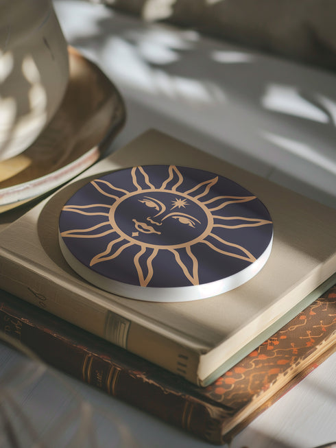 Round ceramic coaster featuring a celestial sun design in warm golden tones against a deep matte black background. The intricate illustration showcases a serene sun face with delicate detailing, surrounded by radiant, curved rays. The smooth, durable surface is complemented by a cork backing for grip and protection, making it both a functional and decorative piece for any space.