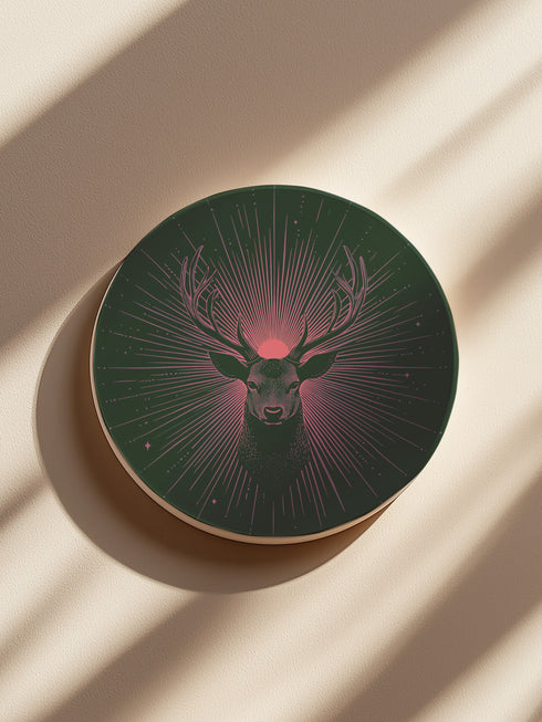 Ceramic coasters in round and square shapes featuring a detailed deer head with majestic antlers against a deep forest green background. A pink-tinged radiant sunburst pattern emanates from behind the deer's head, with delicate stars scattered throughout the design. Cork backing protection.