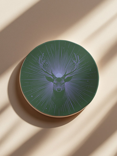 Ceramic coasters in round and square shapes featuring a detailed deer head with antlers on a forest green background, surrounded by radiating periwinkle blue sunburst lines and small stars, with protective cork backing.