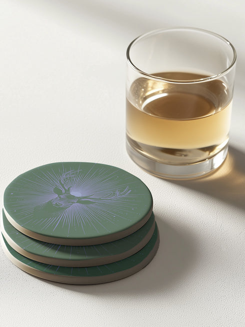 Ceramic coasters in round and square shapes featuring a detailed deer head with antlers on a forest green background, surrounded by radiating periwinkle blue sunburst lines and small stars, with protective cork backing.