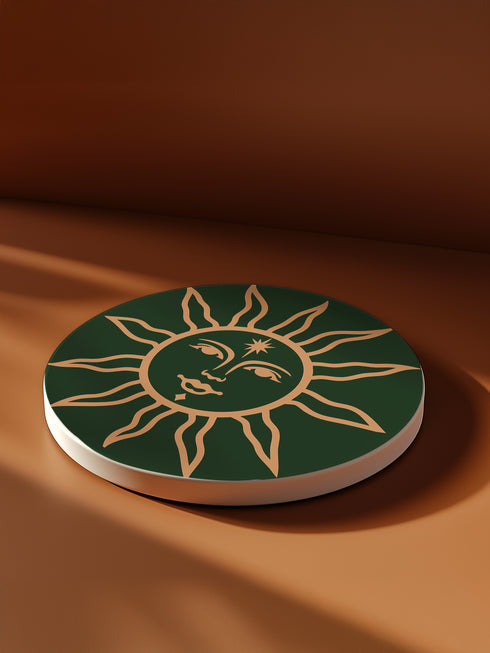 Dark green round ceramic coaster featuring a golden sun illustration with a serene, symmetrical face, delicate curved rays extending outward, and a small star detail on the forehead. The smooth, matte surface enhances the contrast between the deep green background and the warm golden design, giving it a celestial and vintage-inspired aesthetic.