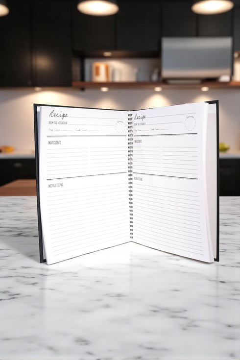 A black spiral-bound hardcover grimoire titled "Spells of Salt & Flame." The cover features an intricate, metallic-effect printed design in silver and gold with an All-Seeing Eye in an archway, flanked by a spoon and an athame. Botanical elements like garlic and herbs grow from a central chalice. The side borders are lined with celestial moon phase symbols. The journal is open, showing blank recipe template pages with fields for ingredients and instructions. The book measures 8.5 x 11 inches.