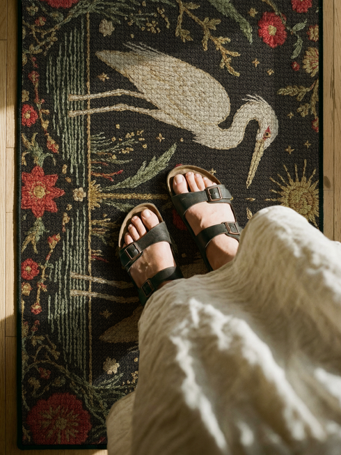 A Grimoire and Goblet rectangular doormat featuring a vintage tapestry design of two white herons facing a central red flower. Above the birds is a golden sun with a face in a celestial style. The background is a dark charcoal with a faux needlepoint texture. The scene is framed by a border of red carnations and green vines in an Arts and Crafts style. The design mimics antique embroidery but is a smooth printed surface with a black trim.