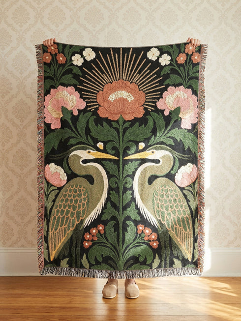 Jacquard woven cotton throw blanket, woven rather than printed, shown flat. Two mirrored great blue herons flank a central radial sunburst, surrounded by botanical blooms and scrollwork. The charcoal black ground carries antique gold ochre sunbeams, blush pink blossoms, deep forest green leaves, muted rust berry clusters, and cream toned herons. A lush multi-colored fringe of black, ochre, and sage threads edges the blanket, and the reverse shows an inverted charcoal grey version of the same design.