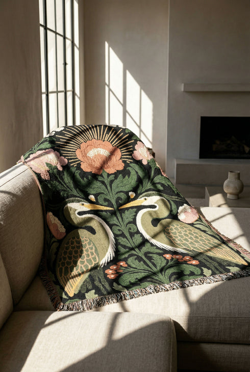 Jacquard woven cotton throw blanket, woven rather than printed, shown flat. Two mirrored great blue herons flank a central radial sunburst, surrounded by botanical blooms and scrollwork. The charcoal black ground carries antique gold ochre sunbeams, blush pink blossoms, deep forest green leaves, muted rust berry clusters, and cream toned herons. A lush multi-colored fringe of black, ochre, and sage threads edges the blanket, and the reverse shows an inverted charcoal grey version of the same design.