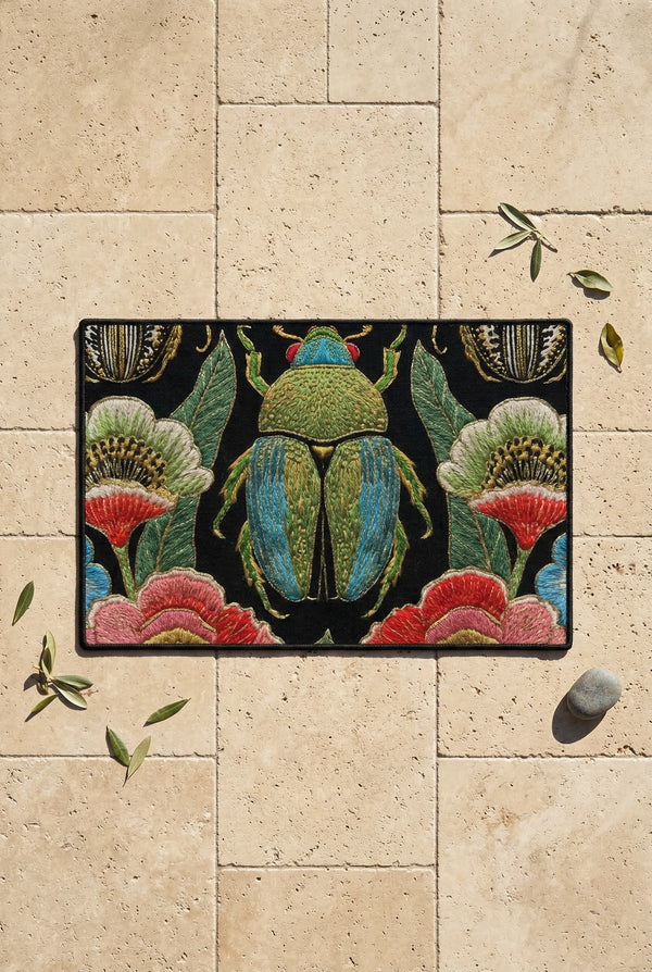 A rectangular doormat featuring a high-fidelity faux embroidery print on a solid black background. The central design is a large, detailed green and teal scarab beetle. It is surrounded by vibrant, flat-printed botanical motifs including large pink, red, and light blue flowers, green foliage, and pale gold-toned seed pods. The mat features a black fabric trim around the edges. The colorful print creates a 2D trompe-l'œil illusion of raised tapestry needlework on a smooth polyester surface.