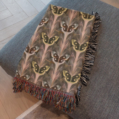 A woven cotton throw featuring an intricate luna moth pattern in bronze and muted earth tones, framed by soft fringe.