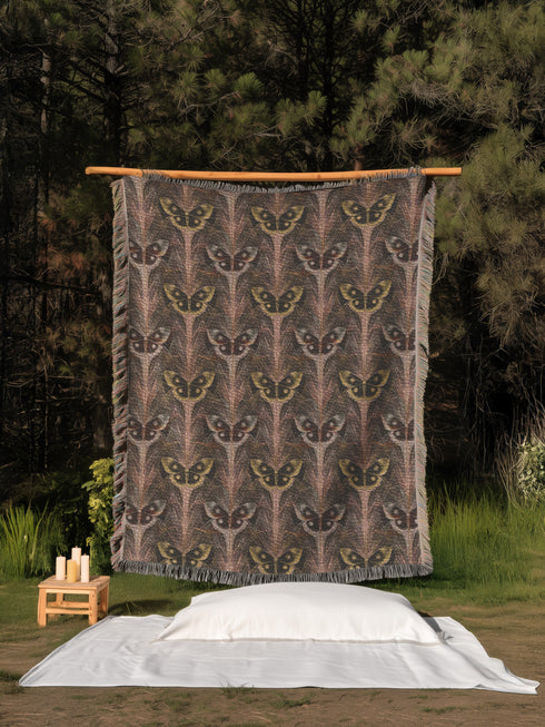 A woven cotton throw featuring an intricate luna moth pattern in bronze and muted earth tones, framed by soft fringe.