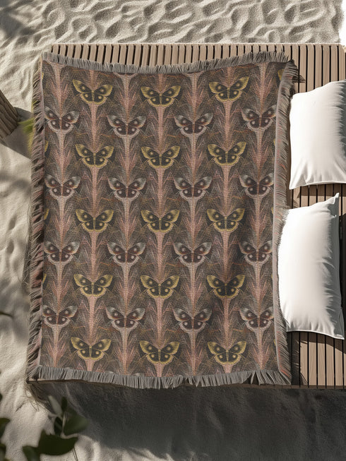 A woven cotton throw featuring an intricate luna moth pattern in bronze and muted earth tones, framed by soft fringe.