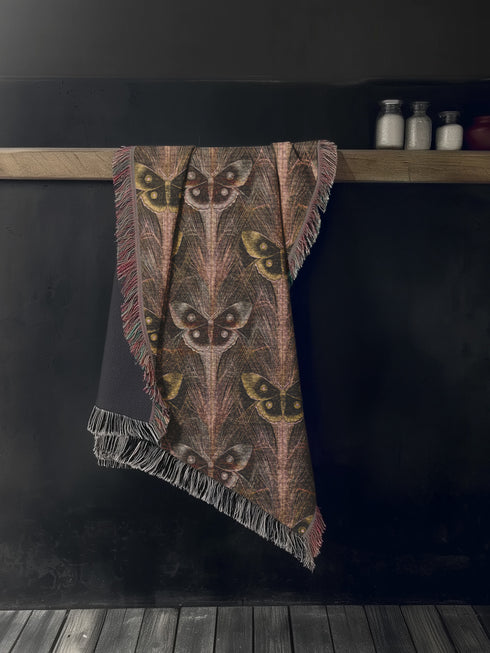 A woven cotton throw featuring an intricate luna moth pattern in bronze and muted earth tones, framed by soft fringe.