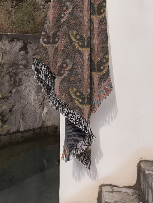 A woven cotton throw featuring an intricate luna moth pattern in bronze and muted earth tones, framed by soft fringe.