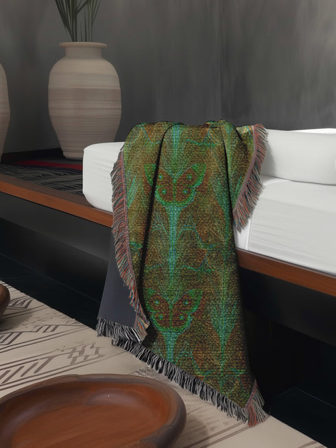 A woven cotton throw featuring an intricate luna moth design in deep green and russet tones, framed by soft fringe.