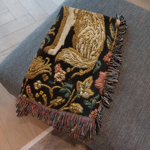 A rectangular, 100% woven cotton throw blanket with a detailed jacquard design. The throw features a symmetrical, vertical pattern of two howling wolves in profile, rendered in shades of beige, gold, and brown against a solid black background. The wolves are surrounded by intricate floral and foliage motifs. The thick, textured weave is visible. The blanket is finished on all four sides with a two-inch, multi-colored fringe that includes black, white, red, and gold threads.