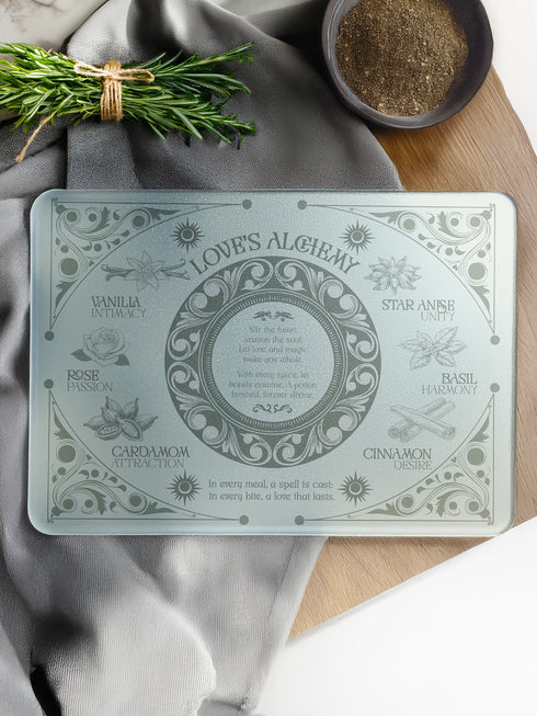 A rectangular sage-tinted glass cutting board featuring an ornate design of magical herbs and spices. Text reads "Love's Alchemy" with detailed illustrations of vanilla, rose, cardamom, star anise, basil, and cinnamon arranged around a central medallion containing a love spell verse.