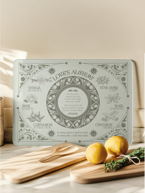 A rectangular sage-tinted glass cutting board featuring an ornate design of magical herbs and spices. Text reads "Love's Alchemy" with detailed illustrations of vanilla, rose, cardamom, star anise, basil, and cinnamon arranged around a central medallion containing a love spell verse.