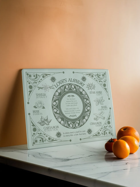 A rectangular sage-tinted glass cutting board featuring an ornate design of magical herbs and spices. Text reads "Love's Alchemy" with detailed illustrations of vanilla, rose, cardamom, star anise, basil, and cinnamon arranged around a central medallion containing a love spell verse.