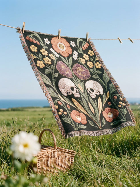 Woven cotton throw blanket with fringed edges and a black ground. Two cream skulls sit side by side at the center of a symmetrical folk garden. A terracotta poppy crowns the top, plum peonies bloom in the middle, and clusters of golden-yellow and white wildflowers, green leaves, and wheat-like sprigs fill the field, with more terracotta poppies along the bottom. The dimensional pattern is woven through a jacquard process, and the reverse shows a mirrored, softer version of the design.