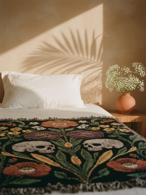Woven cotton throw blanket with fringed edges and a black ground. Two cream skulls sit side by side at the center of a symmetrical folk garden. A terracotta poppy crowns the top, plum peonies bloom in the middle, and clusters of golden-yellow and white wildflowers, green leaves, and wheat-like sprigs fill the field, with more terracotta poppies along the bottom. The dimensional pattern is woven through a jacquard process, and the reverse shows a mirrored, softer version of the design.