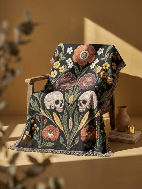 Woven cotton throw blanket with fringed edges and a black ground. Two cream skulls sit side by side at the center of a symmetrical folk garden. A terracotta poppy crowns the top, plum peonies bloom in the middle, and clusters of golden-yellow and white wildflowers, green leaves, and wheat-like sprigs fill the field, with more terracotta poppies along the bottom. The dimensional pattern is woven through a jacquard process, and the reverse shows a mirrored, softer version of the design.
