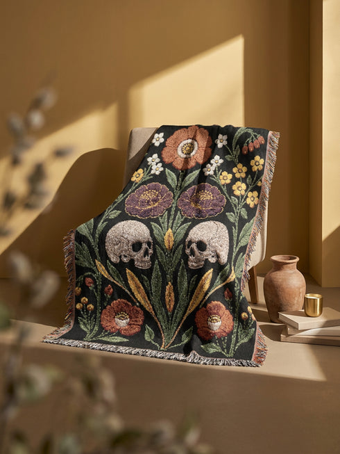 Woven cotton throw blanket with fringed edges and a black ground. Two cream skulls sit side by side at the center of a symmetrical folk garden. A terracotta poppy crowns the top, plum peonies bloom in the middle, and clusters of golden-yellow and white wildflowers, green leaves, and wheat-like sprigs fill the field, with more terracotta poppies along the bottom. The dimensional pattern is woven through a jacquard process, and the reverse shows a mirrored, softer version of the design.
