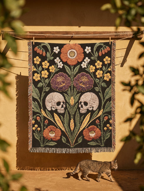 Woven cotton throw blanket with fringed edges and a black ground. Two cream skulls sit side by side at the center of a symmetrical folk garden. A terracotta poppy crowns the top, plum peonies bloom in the middle, and clusters of golden-yellow and white wildflowers, green leaves, and wheat-like sprigs fill the field, with more terracotta poppies along the bottom. The dimensional pattern is woven through a jacquard process, and the reverse shows a mirrored, softer version of the design.
